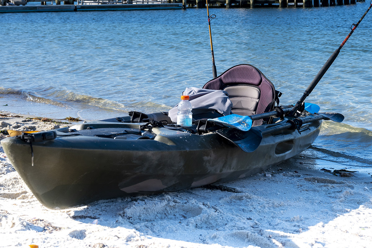 Fishing kayak with equipment stowed on beached on sandy shore with long pier over ocean water in the background